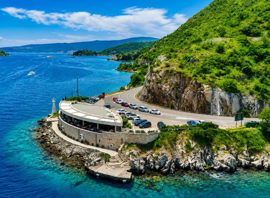 A circular waterfront restaurant on a rocky promontory with parked cars along a curved road overlooking bright blue sea and green hills nearby.