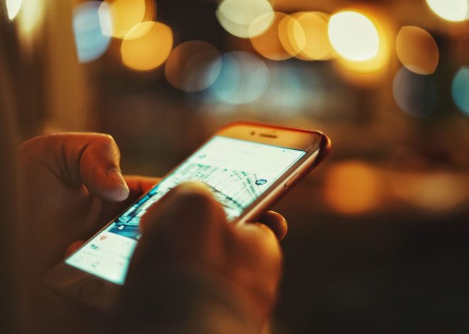 Person holding a smartphone at night, screen glowing while blurred city lights create bokeh in the background.