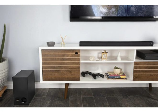 Modern living room media console with a TV mounted above, a soundbar, and a subwoofer on the floor to the left; shelves hold books, decor, and game controllers.
