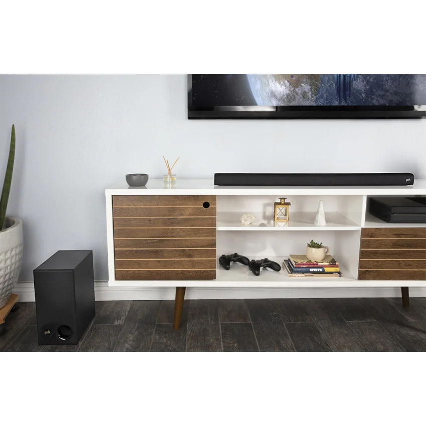 Modern living room media console with a TV mounted above, a soundbar, and a subwoofer on the floor to the left; shelves hold books, decor, and game controllers.