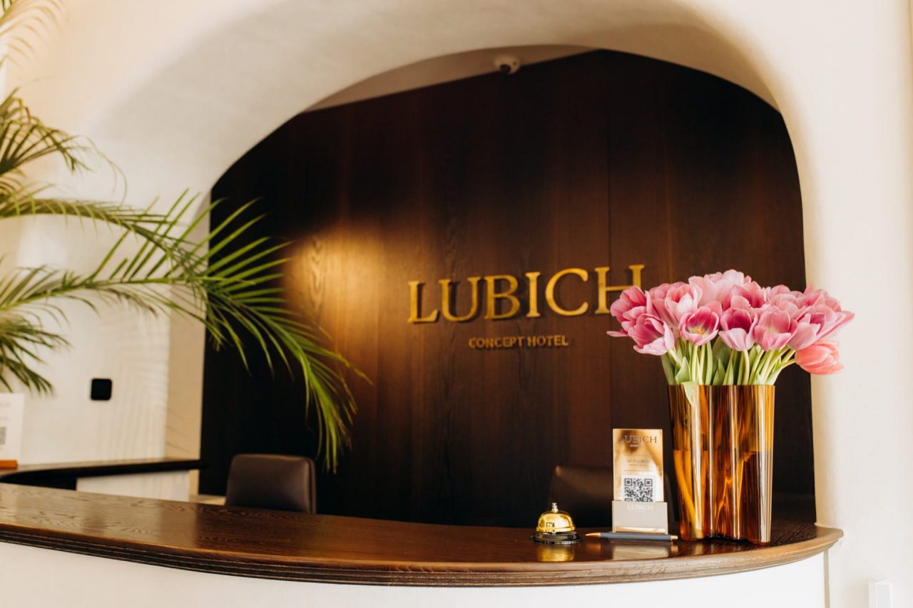 Reception desk at Lubich Concept Hotel with a dark wood backdrop and a gold sign, plus a copper vase of pink tulips on the counter