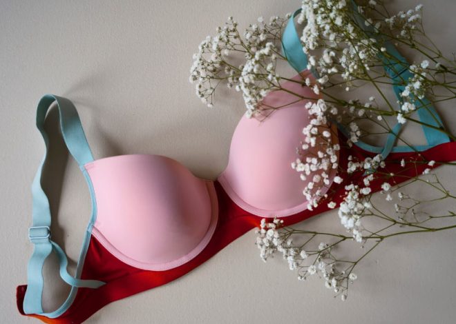 Pink padded bra with red band and light blue straps laid on a surface, accented by sprigs of white baby's breath flowers nearby.