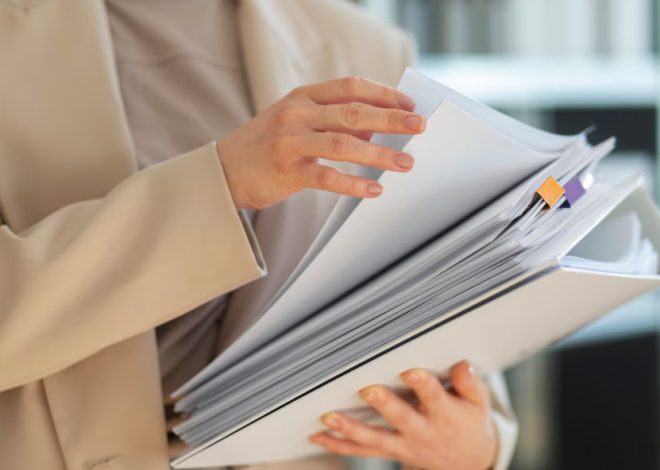 Person in a beige blazer holds a thick binder with many pages and color-coded tabs in an office setting.