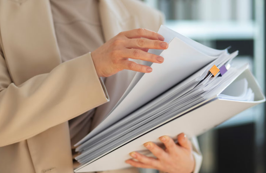 Person in a beige blazer holds a thick binder with many pages and color-coded tabs in an office setting.