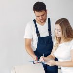 Man in dark blue overalls assists a woman signing a clipboard beside a cardboard box.