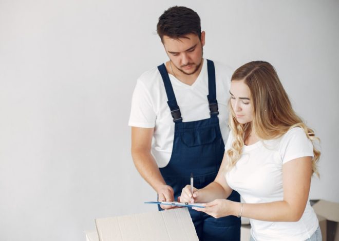 Man in dark blue overalls assists a woman signing a clipboard beside a cardboard box.