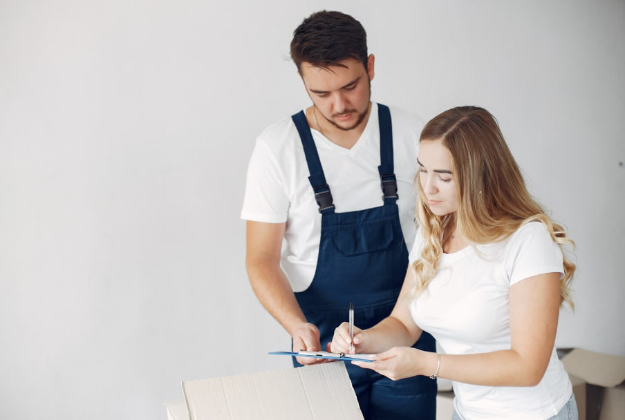 Man in dark blue overalls assists a woman signing a clipboard beside a cardboard box.