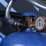 Close-up of blue gas cylinders with brass pressure regulator and round gauge connected to metal piping.