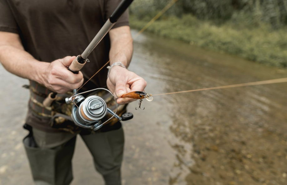 Close-up of a person tying an orange fishing lure to a line on a spinning reel beside a shallow river