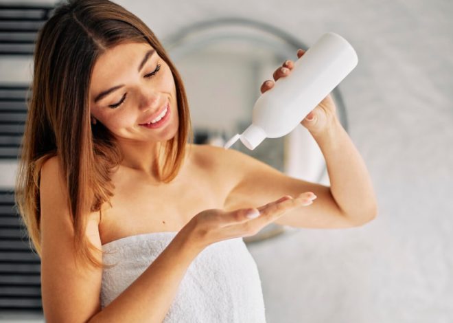 Woman in a towel squeezes lotion from a white bottle into her palm in a bathroom mirrored behind her