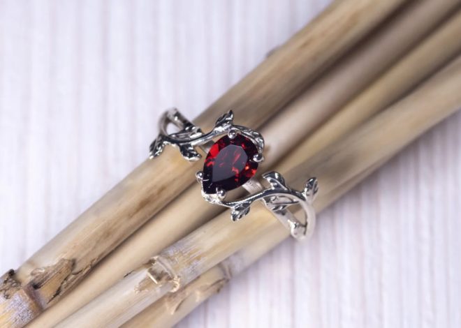 Close-up of a silver ring with a red oval gemstone set on a vine-like band, resting on light wooden sticks.