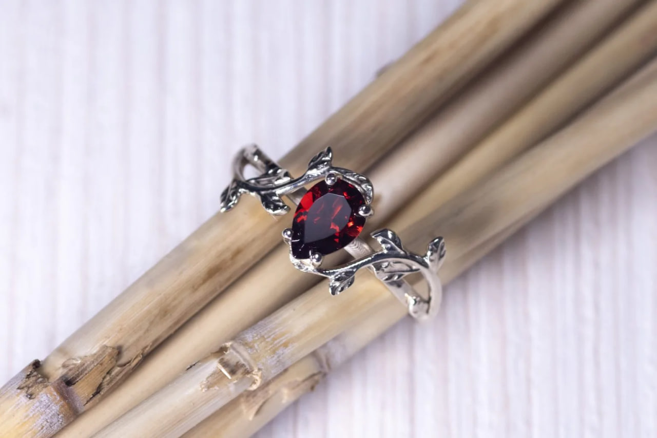 Close-up of a silver ring with a red oval gemstone set on a vine-like band, resting on light wooden sticks.