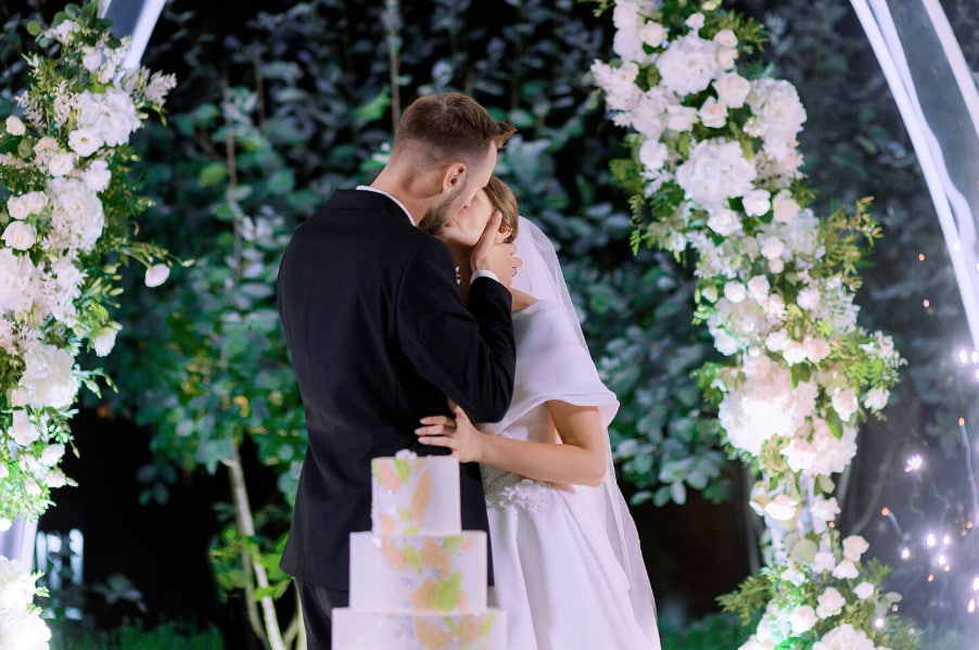 Bride and groom sharing a kiss beneath a white floral arch at night, with a tiered wedding cake in the foreground.