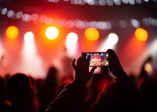Fan in the foreground films a live concert with a smartphone as stage lights blaze in red and white behind them.
