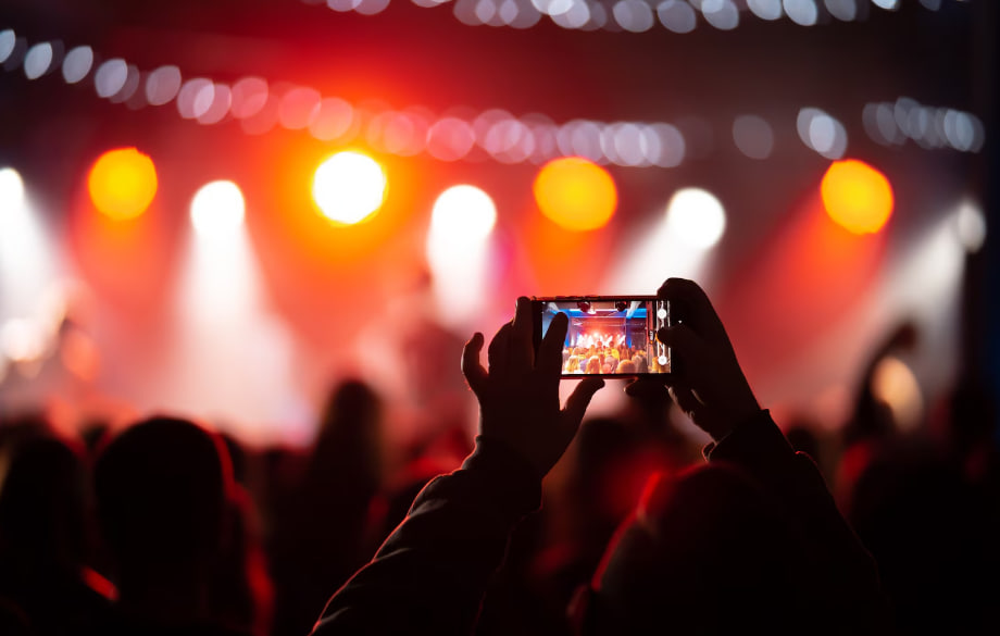 Fan in the foreground films a live concert with a smartphone as stage lights blaze in red and white behind them.