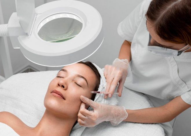 Clinician in gloves administers an injectable facial treatment to a woman under a magnifying lamp in a spa/clinic setting.