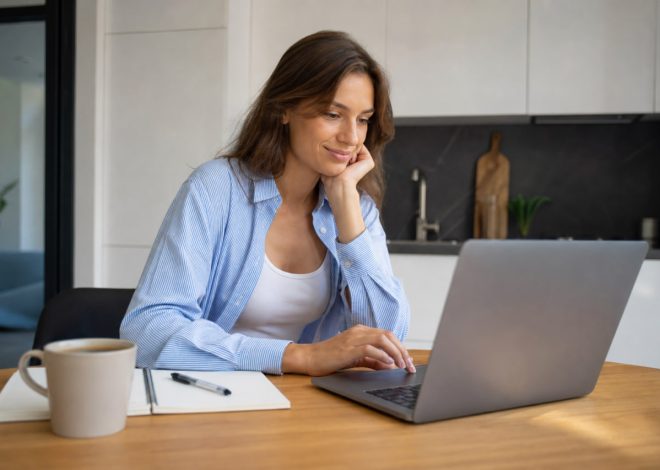 Woman seated at a wooden table typing on a laptop, chin resting on her hand, with a notebook and coffee mug nearby in a modern kitchen setting.