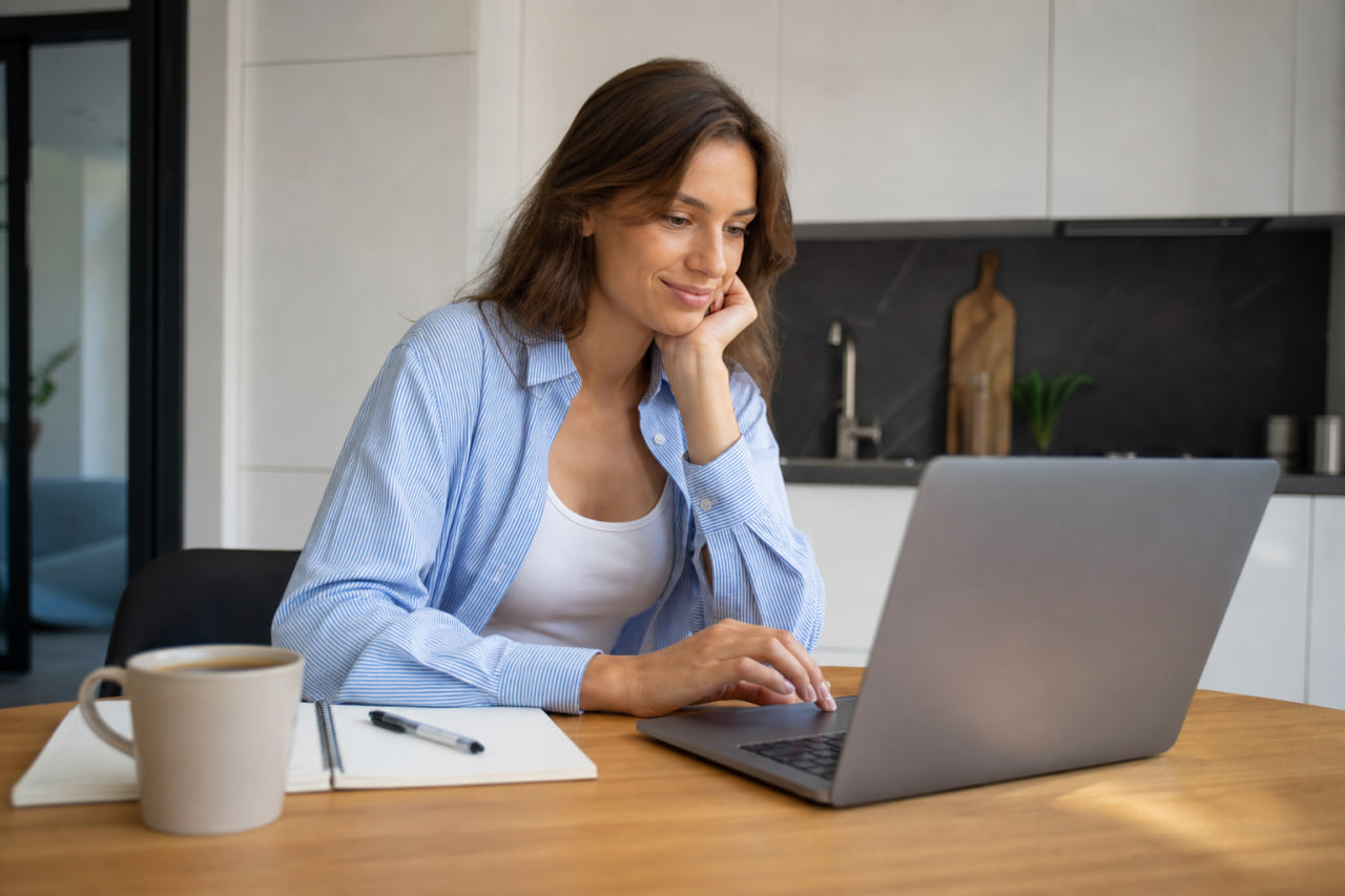 Woman seated at a wooden table typing on a laptop, chin resting on her hand, with a notebook and coffee mug nearby in a modern kitchen setting.