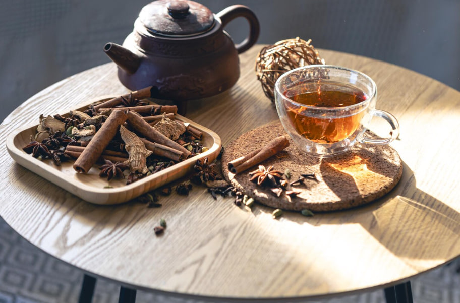 Glass cup of tea on a cork coaster with a brown teapot and a tray of cinnamon sticks and spices on a wooden table.
