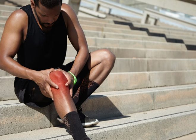 Athlete sits on outdoor concrete steps, holding a reddened knee with a worried expression, suggesting an injury.