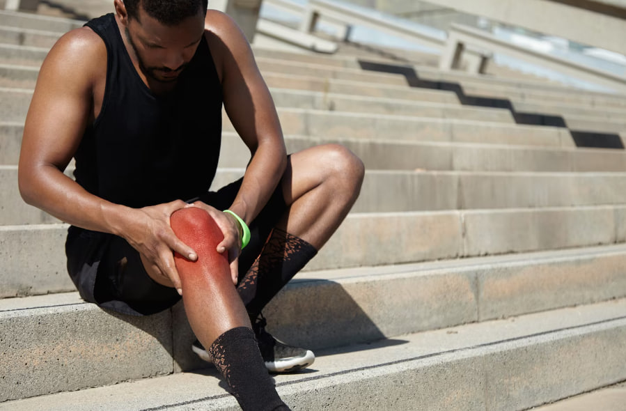 Athlete sits on outdoor concrete steps, holding a reddened knee with a worried expression, suggesting an injury.