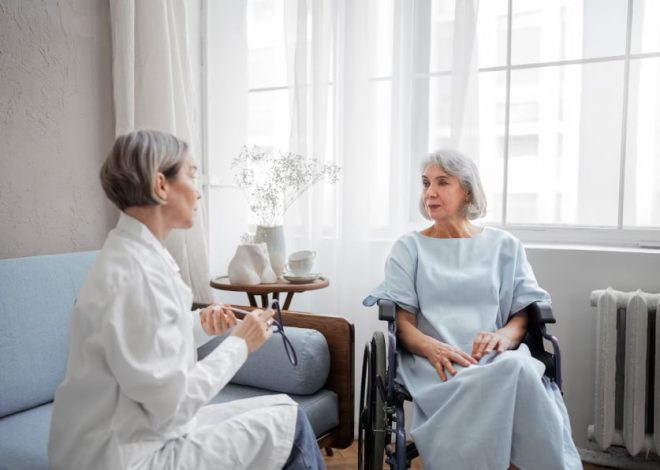 Healthcare professional in a white coat speaks with an elderly woman in a wheelchair in a bright patient room by a small table with a vase.