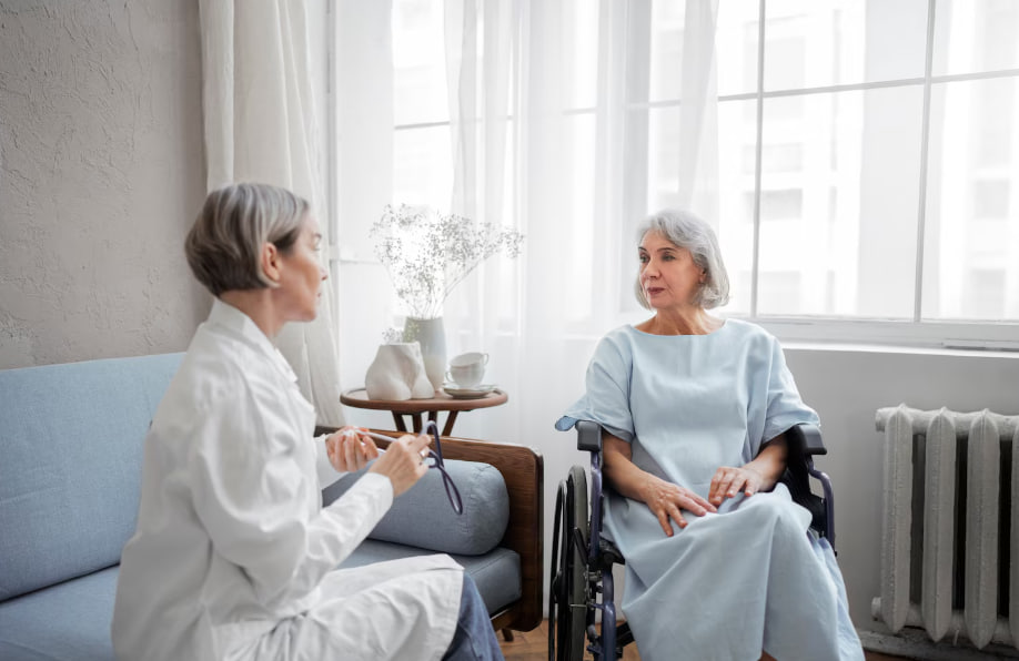 Healthcare professional in a white coat speaks with an elderly woman in a wheelchair in a bright patient room by a small table with a vase.