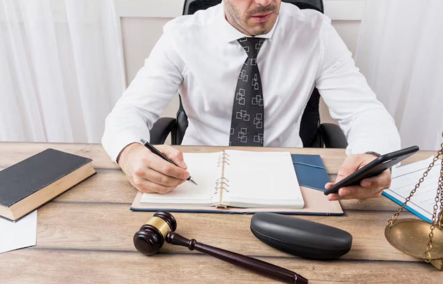 Lawyer at a desk writing in a notebook while holding a smartphone, with a gavel and scales nearby.