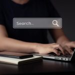 Person typing on a laptop at a dark desk; a translucent search box reading 'Search...' floats above.