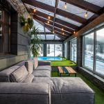 Cozy sunroom with a gray sectional sofa, wooden coffee table, and string lights, overlooking a snowy yard and blue pool outside through large windows.