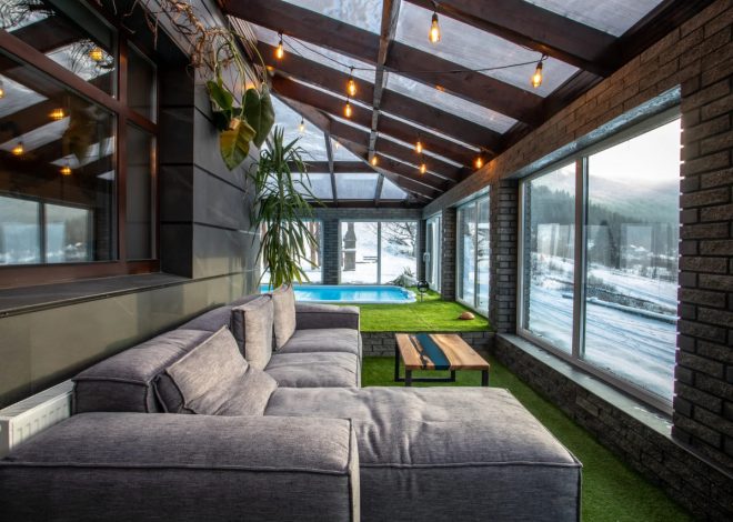 Cozy sunroom with a gray sectional sofa, wooden coffee table, and string lights, overlooking a snowy yard and blue pool outside through large windows.