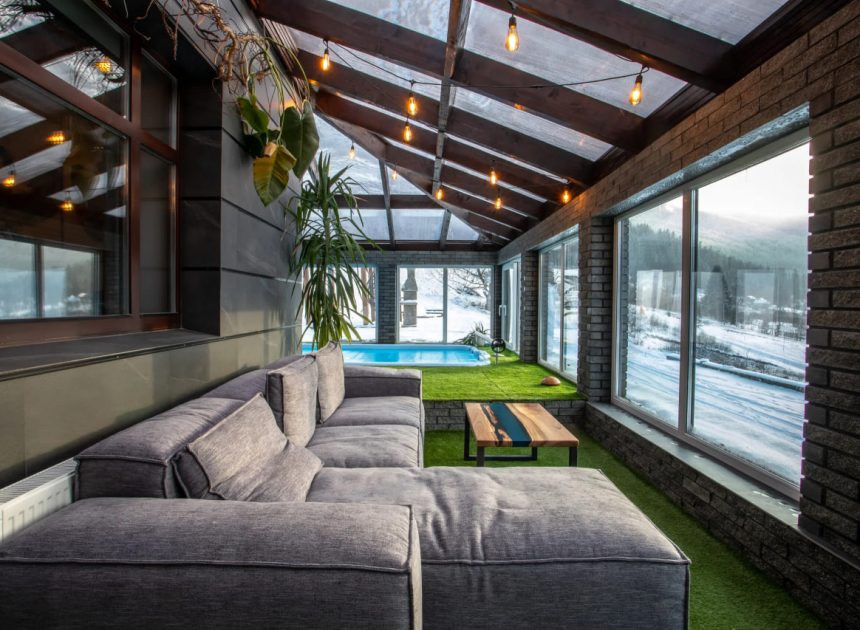 Cozy sunroom with a gray sectional sofa, wooden coffee table, and string lights, overlooking a snowy yard and blue pool outside through large windows.