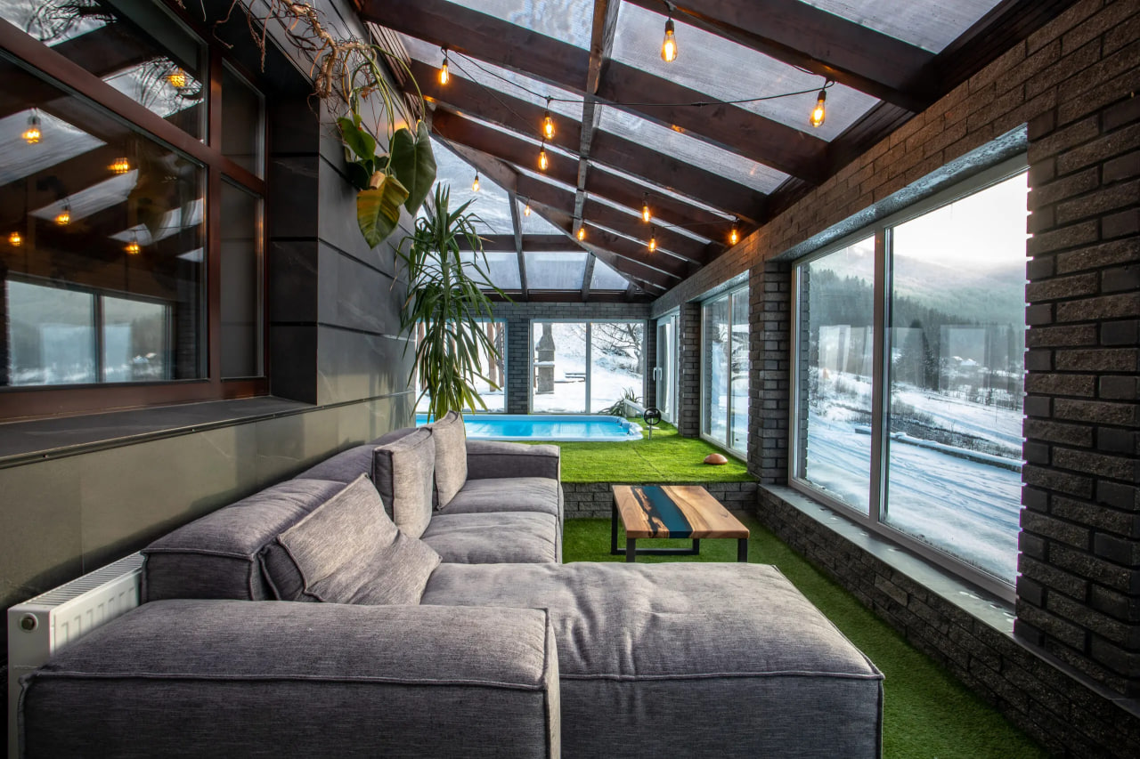 Cozy sunroom with a gray sectional sofa, wooden coffee table, and string lights, overlooking a snowy yard and blue pool outside through large windows.