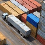 A truck with a long trailer drives between rows of stacked shipping containers in a busy yard.