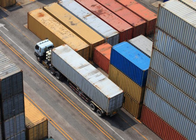 A truck with a long trailer drives between rows of stacked shipping containers in a busy yard.