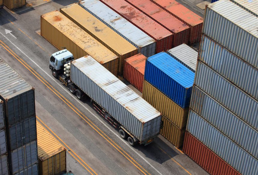 A truck with a long trailer drives between rows of stacked shipping containers in a busy yard.