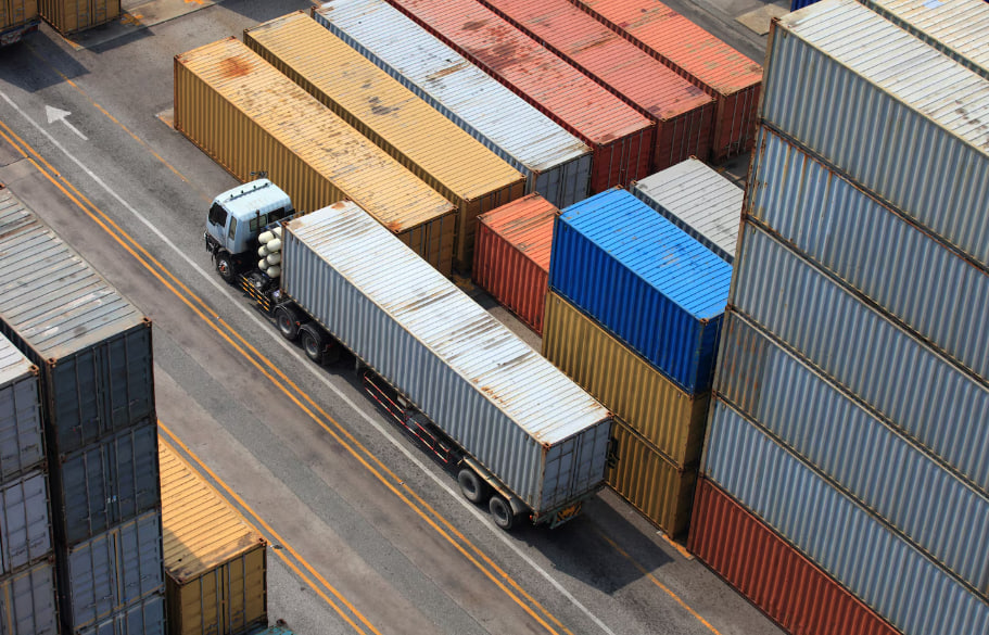 A truck with a long trailer drives between rows of stacked shipping containers in a busy yard.