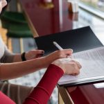 Two people sign or review a document at a glossy desk by a window, one using a pen on the pages.