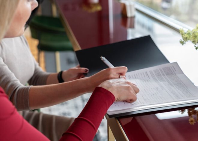 Two people sign or review a document at a glossy desk by a window, one using a pen on the pages.