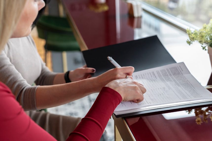Two people sign or review a document at a glossy desk by a window, one using a pen on the pages.
