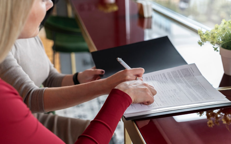 Two people sign or review a document at a glossy desk by a window, one using a pen on the pages.