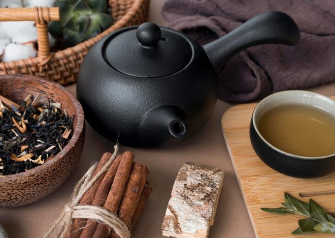 Black teapot with a cup of tea, cinnamon sticks tied with twine, and a bowl of loose leaf tea on a wooden surface.