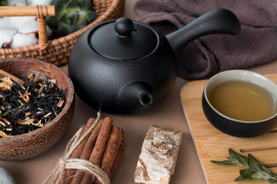 Black teapot with a cup of tea, cinnamon sticks tied with twine, and a bowl of loose leaf tea on a wooden surface.