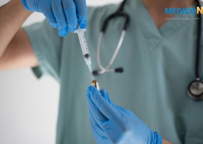 Healthcare professional in blue gloves draws liquid from a vial with a syringe; stethoscope in view on scrubs.