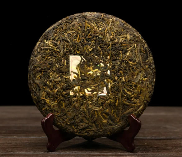 Round compressed tea cake made of dark leaves on a wooden stand against a black background.