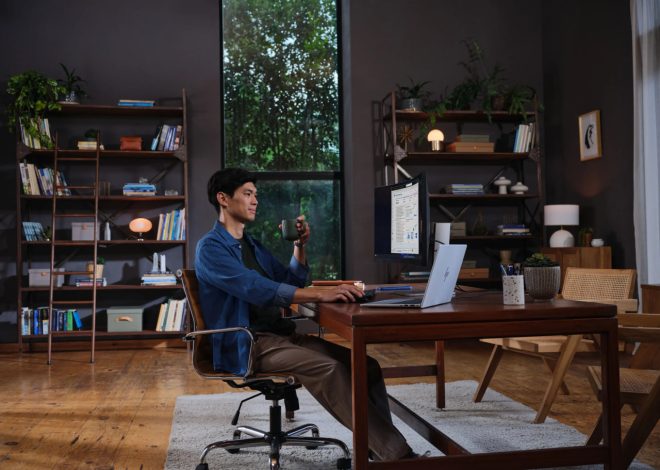 Man sits at a wooden desk in a home office, using a laptop with an external monitor and holding a mug.