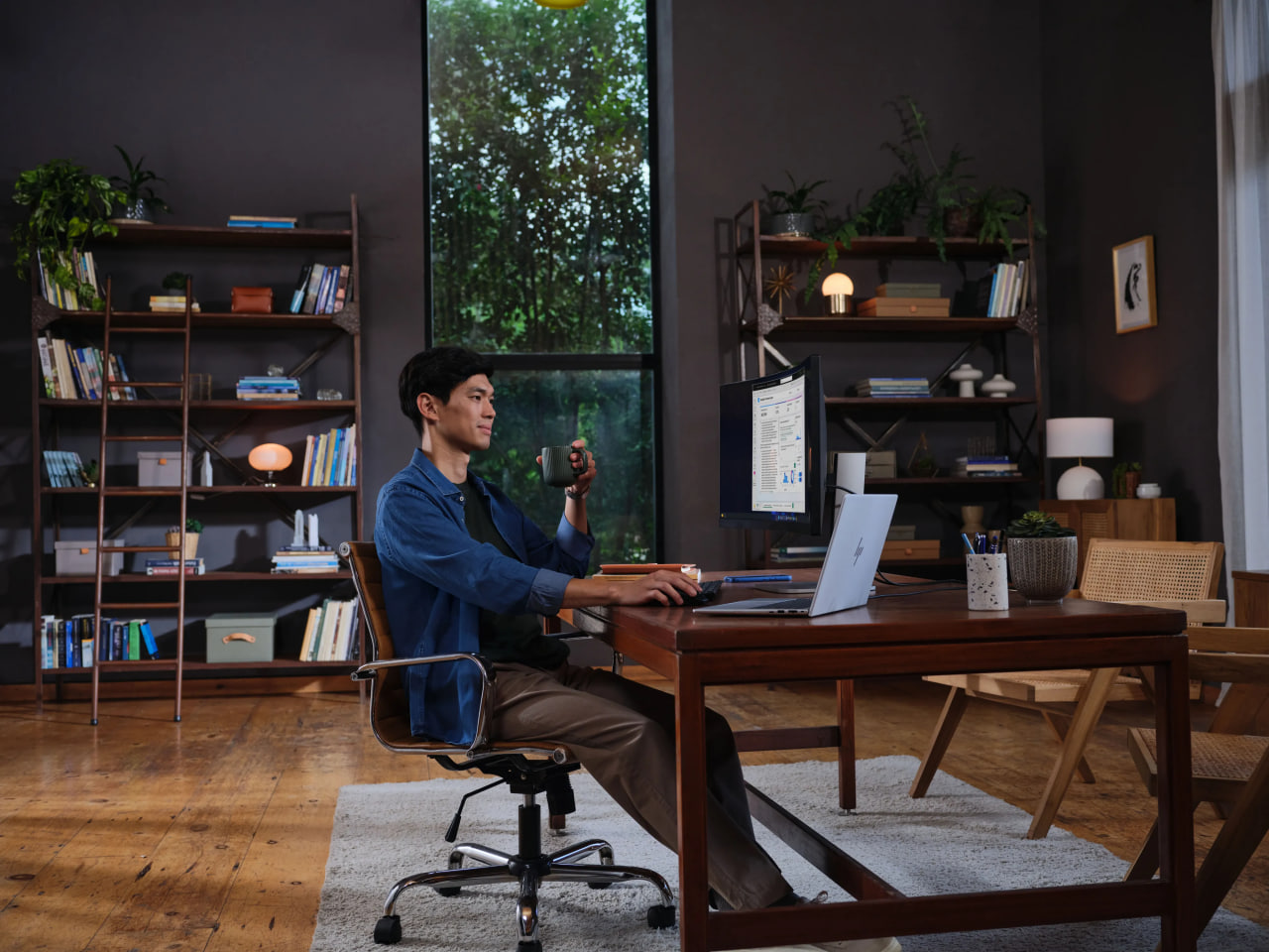 Man sits at a wooden desk in a home office, using a laptop with an external monitor and holding a mug.