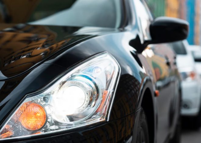 Close-up of a black car's headlight and front fender with reflections on its glossy surface in an urban street setting.