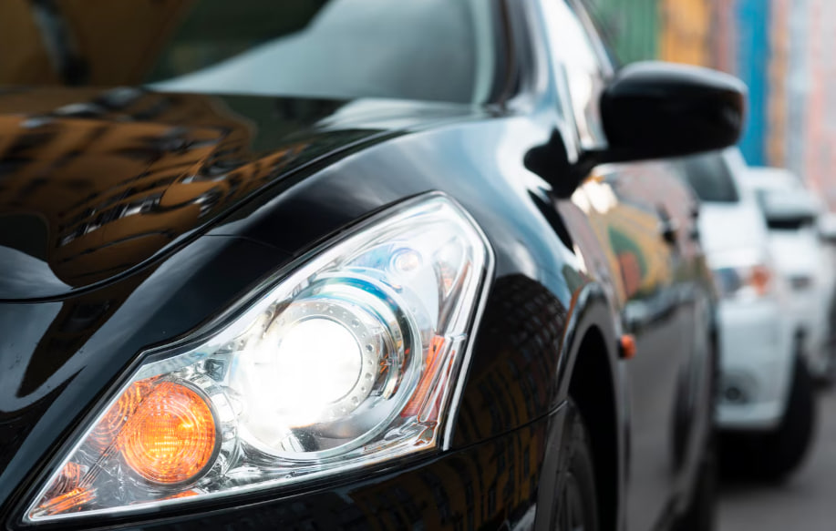 Close-up of a black car's headlight and front fender with reflections on its glossy surface in an urban street setting.