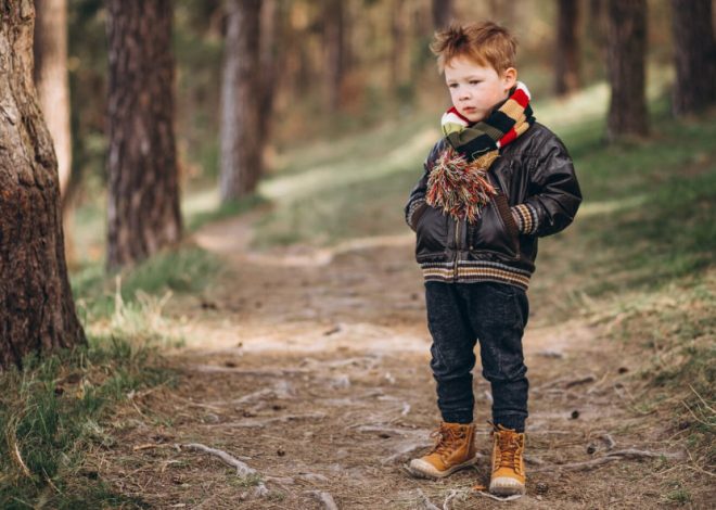 Young boy in a forest path, wearing a leather jacket, striped scarf, and tan boots, holding a colorful pompom.